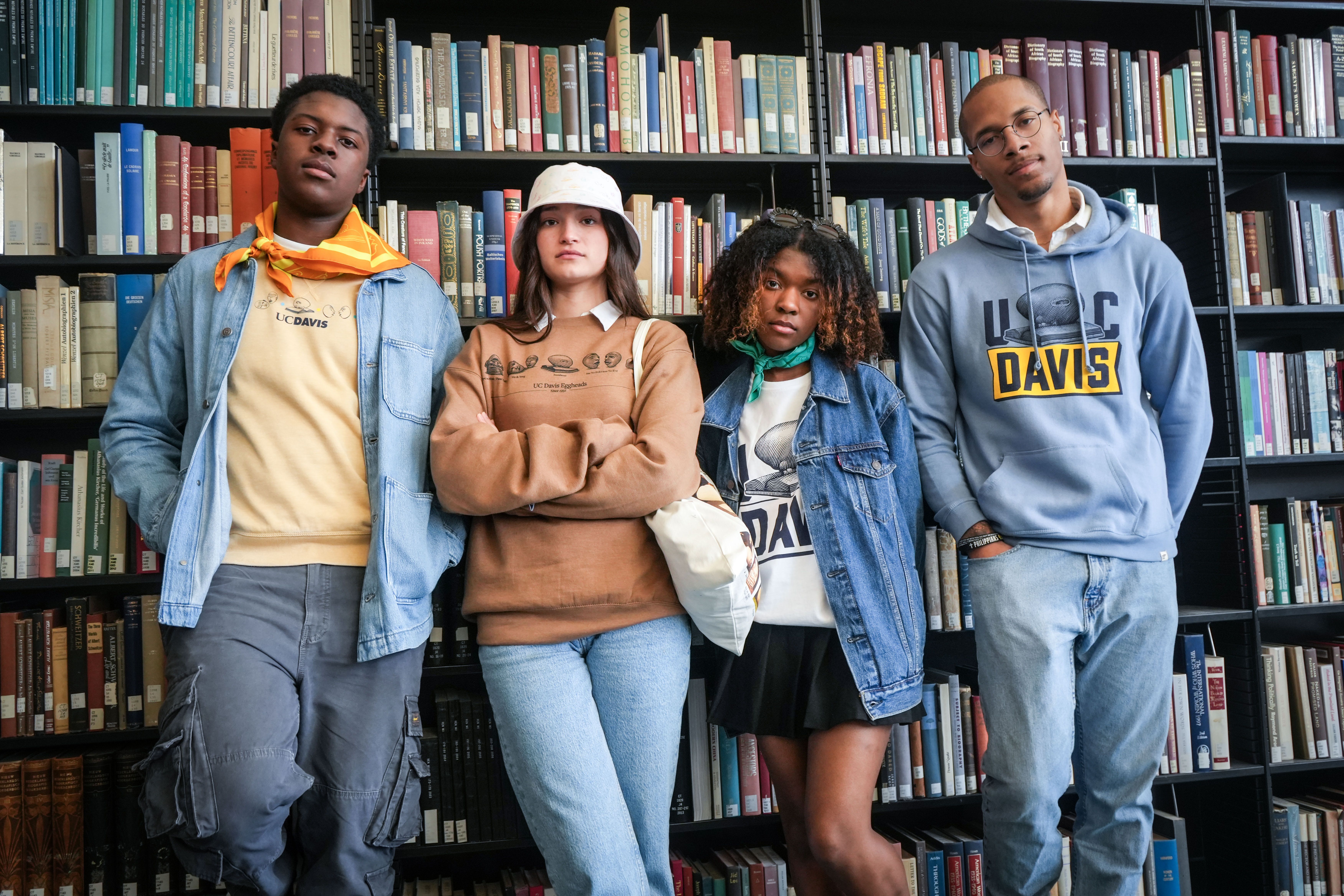 four students stand next to each other and face the camera in a library setting. all four are wearing apparel from the new Arneson Egghead Collection featuring the Egghead sculptures made by former ˽̳ Davis faculty Robert Arneson.