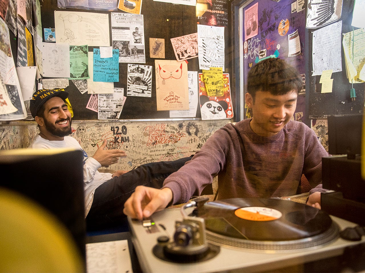 A ˽̳ Davis student works a record player in a radio studio while another student sits in front of the poster-filled back wall, smiling.