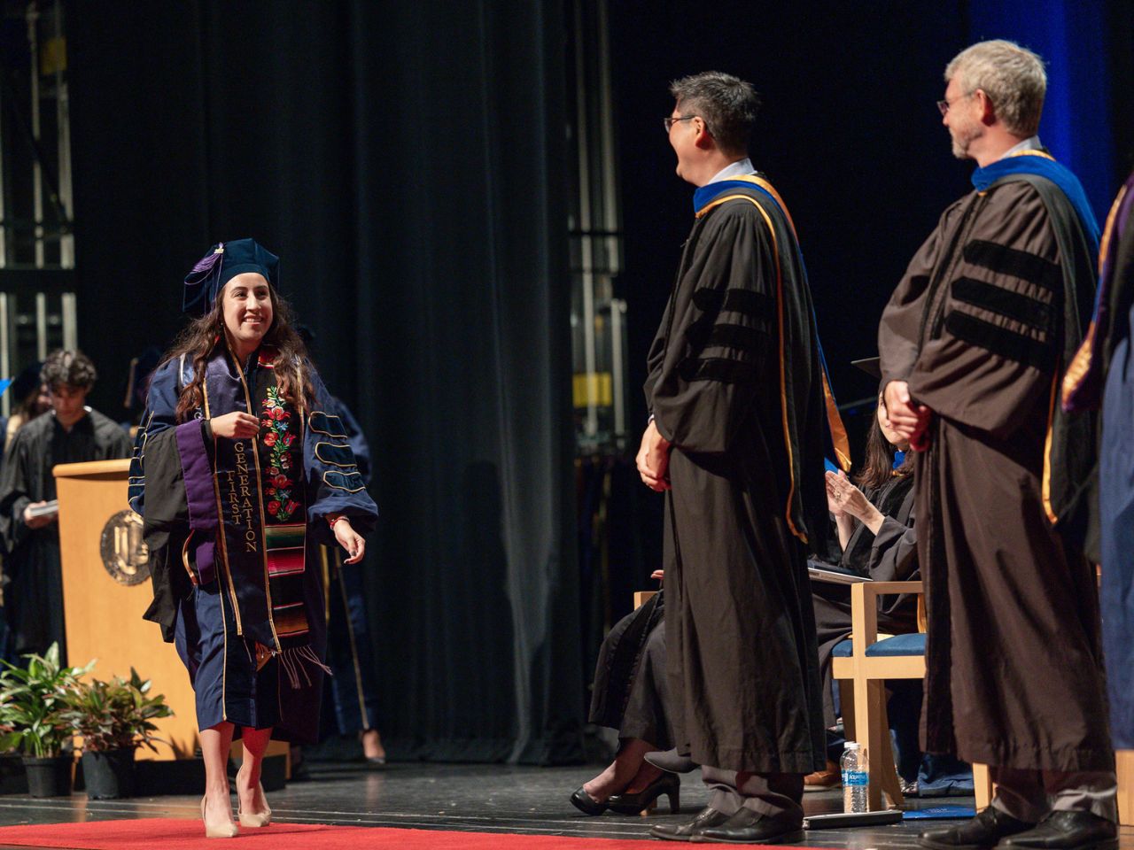 A student in a blue commencement gown and regalia, including a stole with the words "First Generation" walks across the stage at the ˽̳ Davis School of Law commencement ceremony.