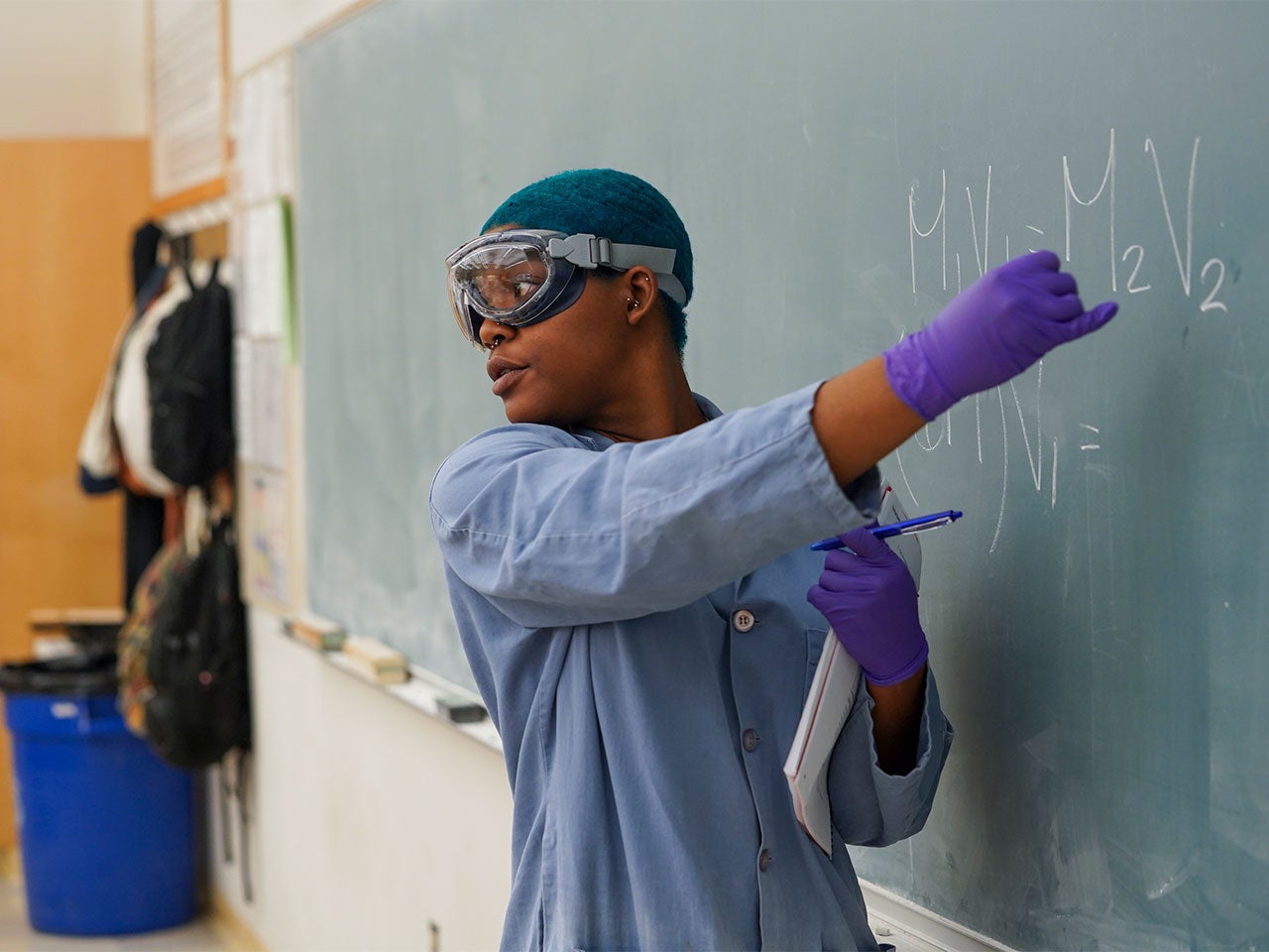 A ˽̳ Davis teaching assistant with short blue hair and wearing a blue lab coat points to a chemical equation on a blackboard. 