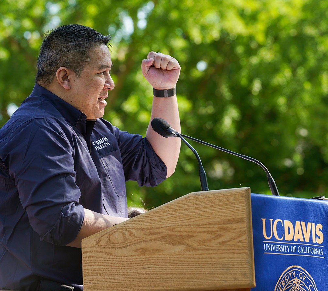 Dr. Nam Tran, in a ˽̳ Davis Health shirt, speaks at a podium with his fist raised at Picnic Day's opening ceremony, celebrating his role as both alum and 2022 Parade Marshal.