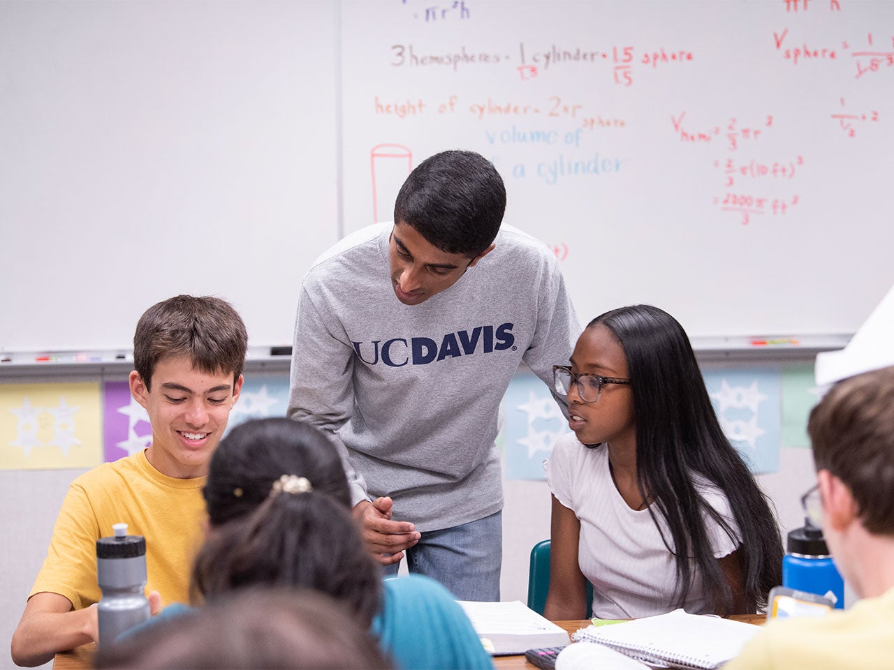 A student wearing a light gray long-sleeve ˽̳ Davis shirt leans down to assist two junior high school students. In the background is a large whiteboard with equations written on it.