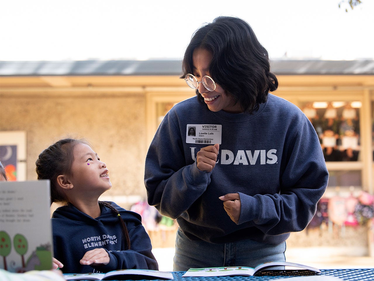 A student in a dark blue ˽̳ Davis sweater and round-rimmed glasses helps an elementary student read a picture book. The elementary student looks up at the ˽̳ Davis student with a big smile.