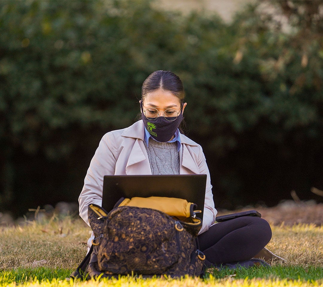Student working on a laptop outdoors at ˽̳ Davis Arboretum, maintaining health protocols with a face mask.