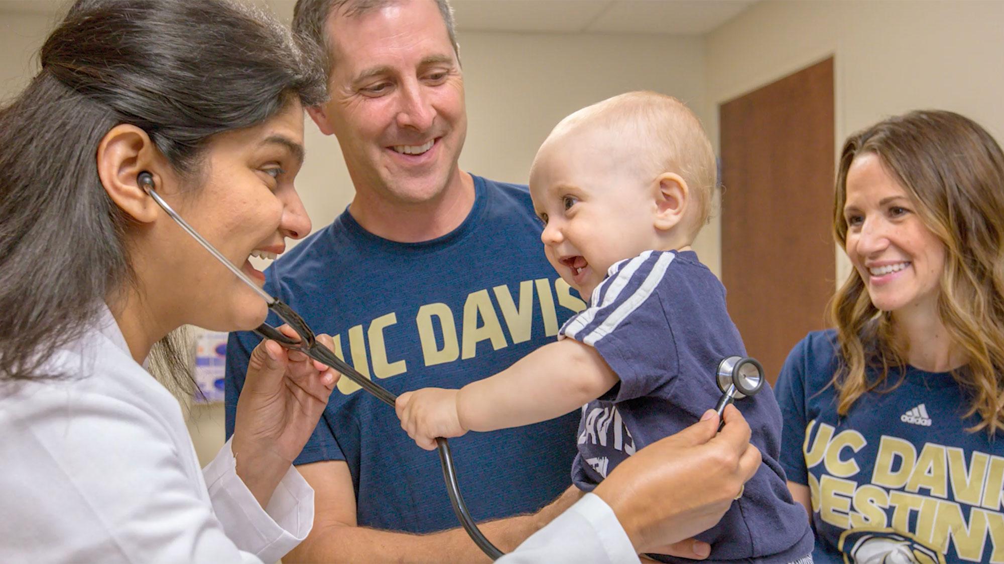 Two parents dressed in ˽̳ Davis Aggie gear present their baby to a doctor at a checkup