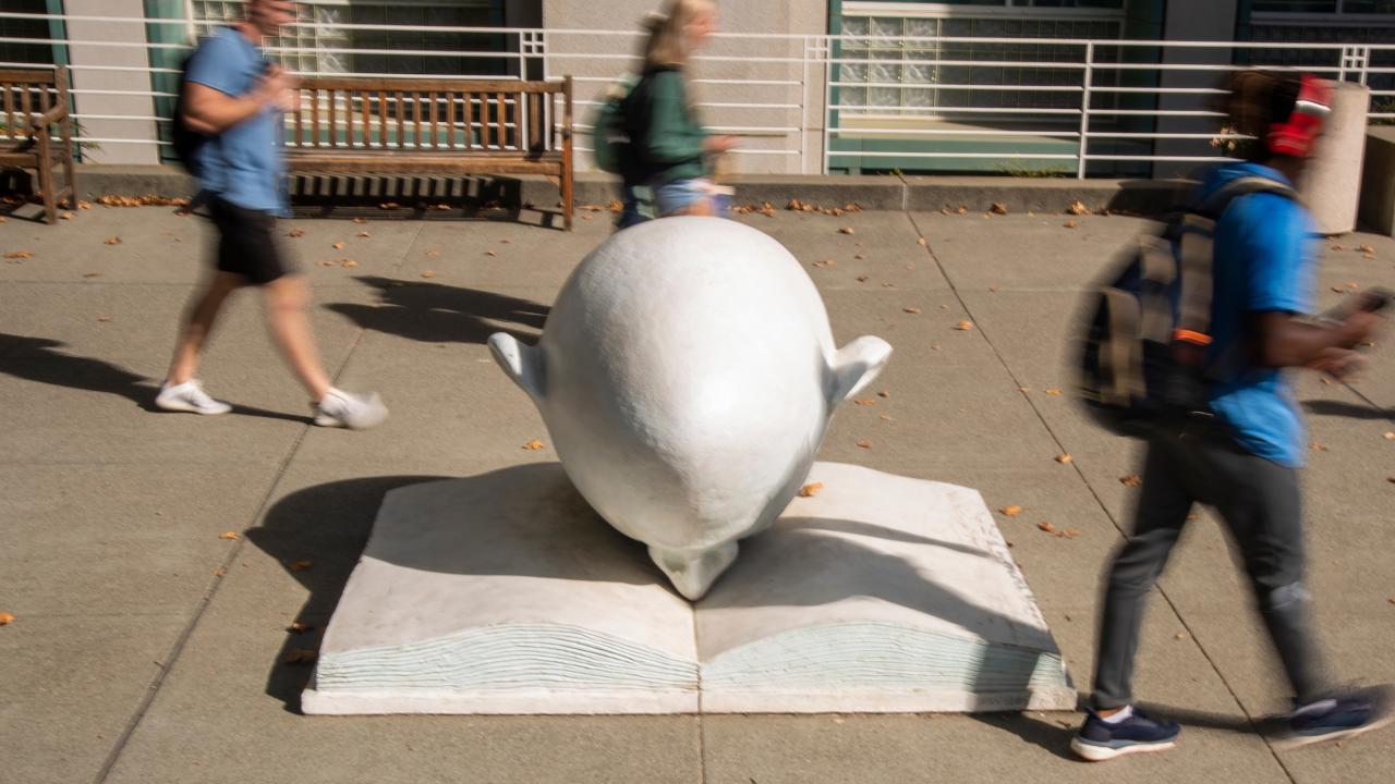 Bookhead Egghead shot in front of library at ˽̳ Davis with students, in blur, walking by