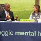 ˽̳ Davis Chancellor Gary S. May in suit, sits at table, chatting with two Aggie Mental Health Ambassadors are tabling, on the Quad